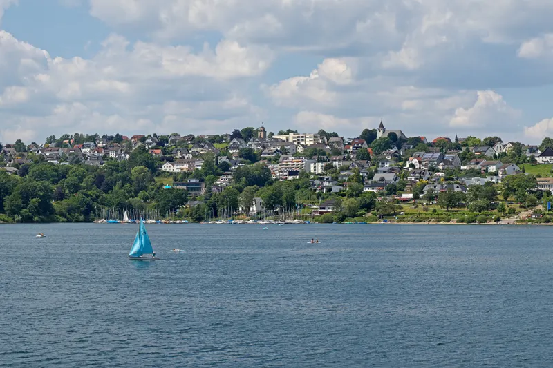 BookNoFees: Langscheid am Sorpesee bei Sundern: das Dorf am Seeufer hoch über dem Wasser, mit einem Segelboot im Vordergrund