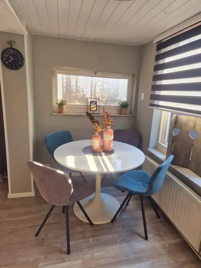 BookNoFees: Ferienwohnung in Domburg – Essbereich mit rundem weißem Tisch, blauen und grauen Stühlen und einem Fenster mit Blick auf den Wald